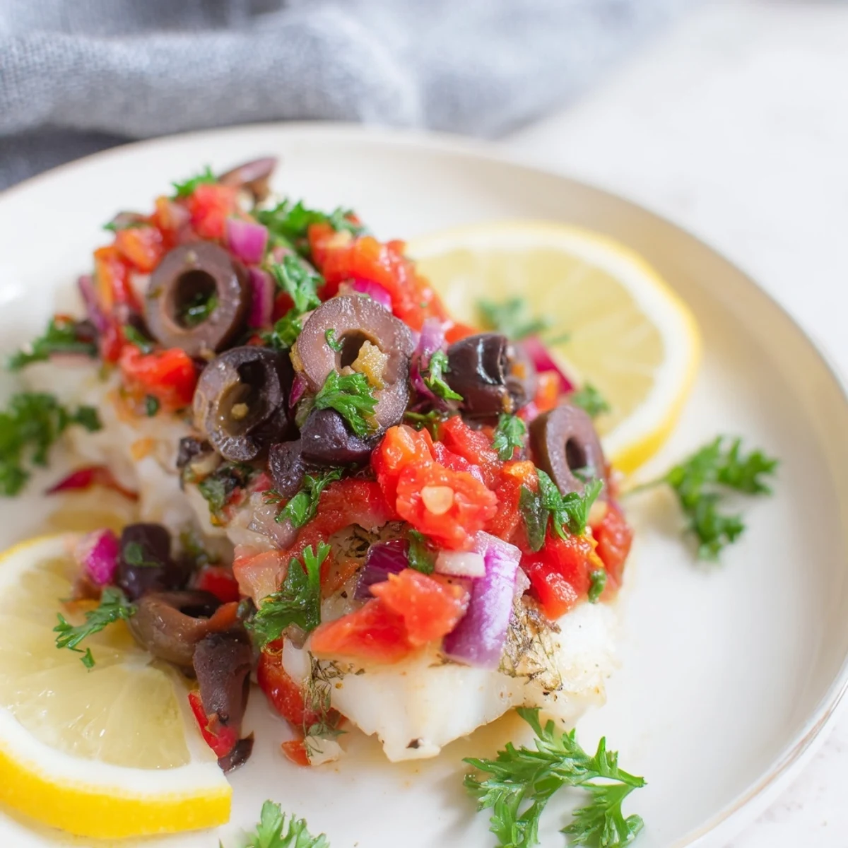 A close-up of baked cod with tomato and olive salsa, flaky white fish garnished with fresh parsley and capers.