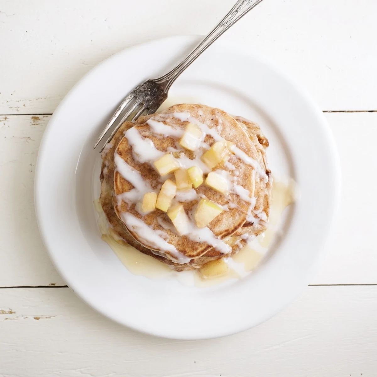 Stack of golden brown Apple Fritter Pancakes drizzled with white glaze, served on a rustic plate with a side of warm maple syrup.  
