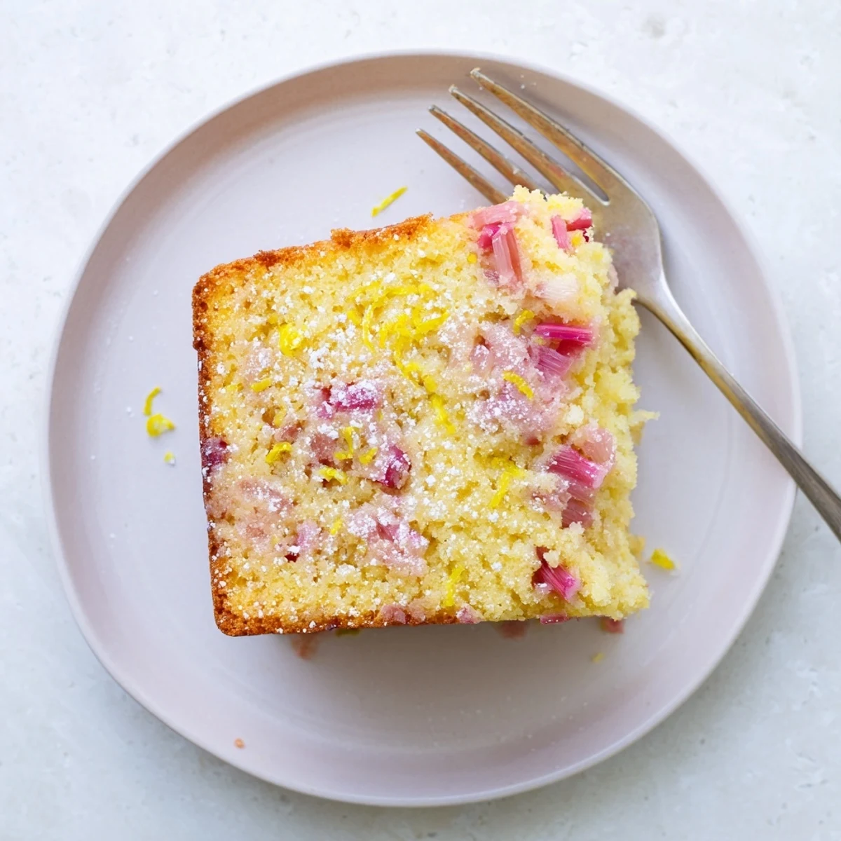 Moist zesty rhubarb lemon drizzle cake serving with tart pink rhubarb chunks throughout the golden crumb