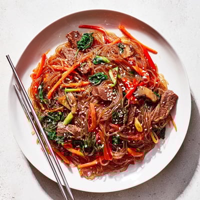 Steam rises from a bowl of Korean Beef Noodles, showing springy glass noodles and wilted spinach topped with sesame seeds.